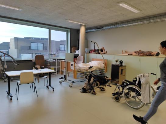 Een vrouw wandelt binnen in een verzorgingslokaal. Hierin staat een ziekenbed, een rolstoel en liggen twee levensechte poppen. Er ligt ook een pop op de grond naast een omgevallen rolstoel.